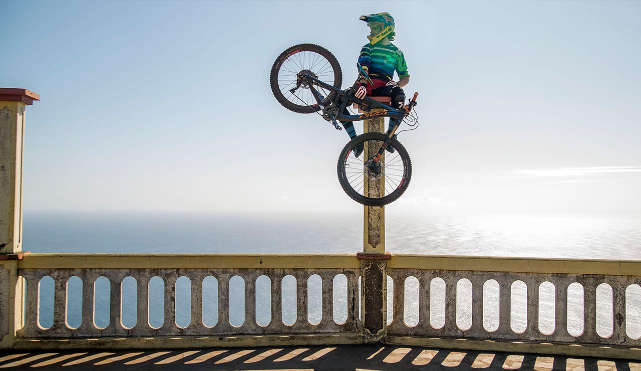 Bam Hill‘s sitting on a large pillar, bike in hand, overlooking the ocean.